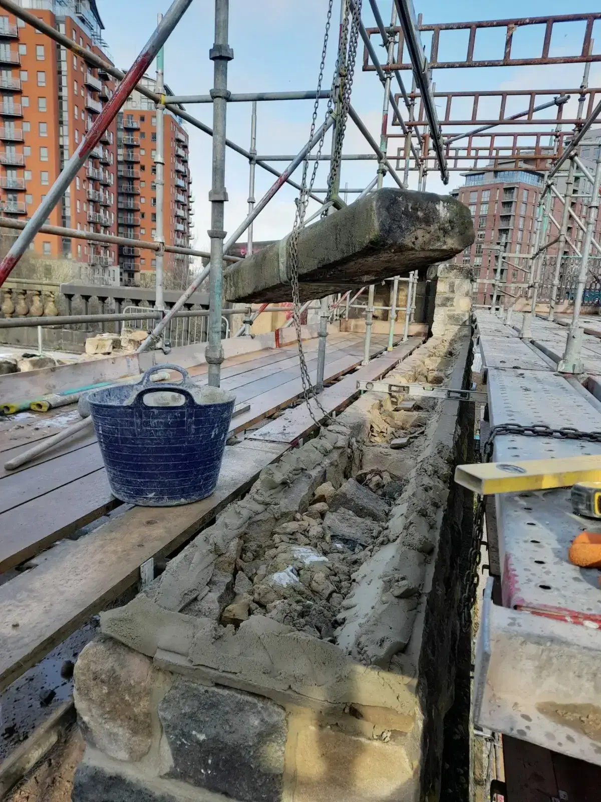 Stone wall restoration in progress on scaffolding in a city setting, with suspended stone lintel and visible masonry work.